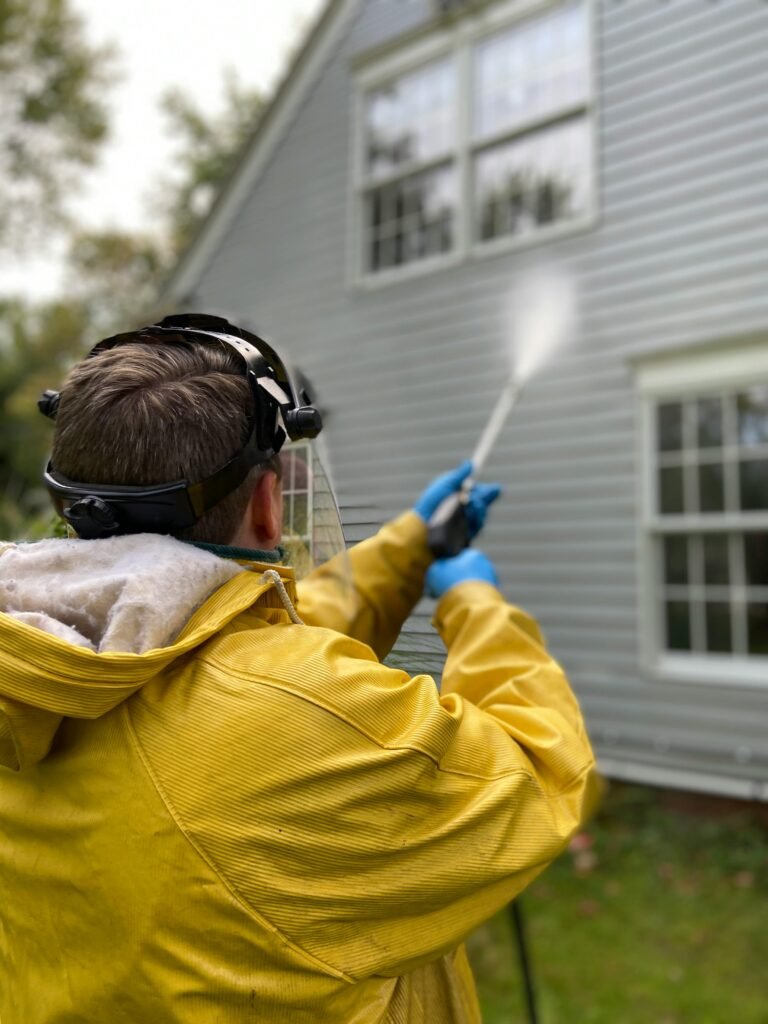 Person cleaning house siding with a pressure washer, wearing protective gear in a yellow raincoat.